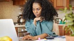 一个年轻的黑人妇女坐在厨房的桌子旁，文件散乱，笔记本电脑开着